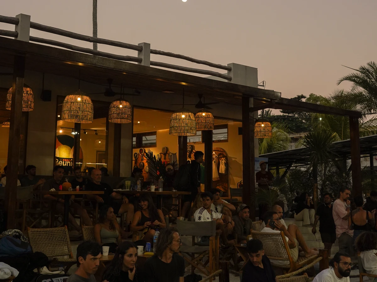 A crowd relaxing in lounge chairs under woven lamps at dusk, the moon above the clubhouse